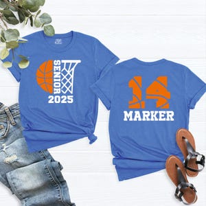 May include: Two blue t-shirts with basketball-themed designs. The first shirt has a basketball and hoop with the text "Senior 2025". The second shirt has a basketball number "14" and the text "Marker".