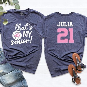 May include: Two navy blue t-shirts. The front shirt has a white volleyball with the number 21 and the text "that's my senior!" The back shirt has the name "JULIA" and the number "21" in pink.