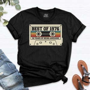 May include: A black short-sleeve t-shirt featuring a graphic of a cassette tape with the text "BEST OF 1976" and "50 YEARS OF BEING AWESOME" in a retro color palette of orange, yellow, and teal stripes on the front center.