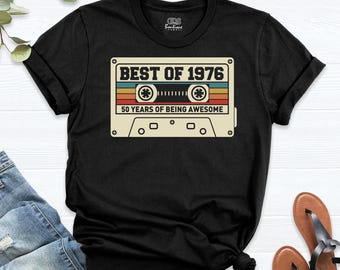 May include: A black short-sleeve t-shirt featuring a graphic of a cassette tape with the text "BEST OF 1976" and "50 YEARS OF BEING AWESOME" in a retro color palette of orange, yellow, and teal stripes on the front center.