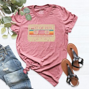 May include: A maroon t-shirt with a retro cassette tape design. The tape graphic features the text "BEST OF 1975" and "50 YEARS OF BEING AWESOME" in a vintage style. The shirt is paired with blue jeans and sandals.