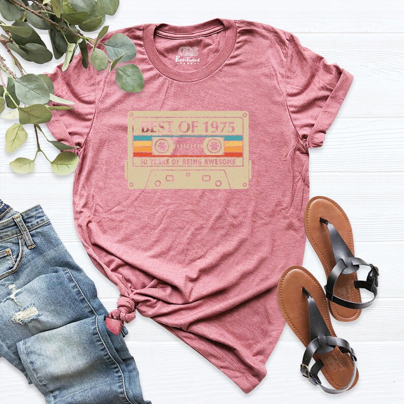 May include: A maroon t-shirt with a retro cassette tape design. The tape graphic features the text "BEST OF 1975" and "50 YEARS OF BEING AWESOME" in a vintage style. The shirt is paired with blue jeans and sandals.