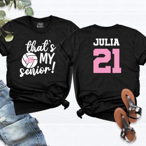 May include: Two black t-shirts with white text. One shirt says "that's my senior!" with a volleyball graphic. The other shirt says "JULIA" with the number "21" in pink.