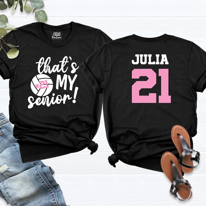 May include: Two black t-shirts with white text. One shirt says "that's my senior!" with a volleyball graphic. The other shirt says "JULIA" with the number "21" in pink.
