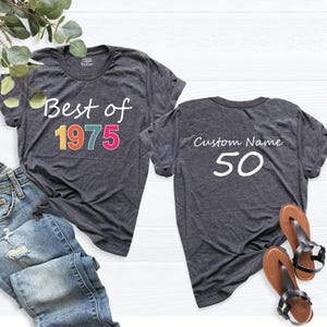 May include: Two gray t-shirts. The front of one shirt says "Best of 1975" in colorful letters. The back of the other shirt says "Custom Name 50".