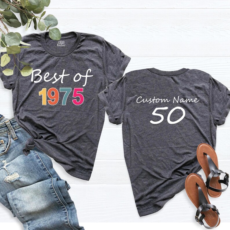 May include: Two gray t-shirts. The front of one shirt says "Best of 1975" in colorful letters. The back of the other shirt says "Custom Name 50".