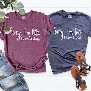 May include: A maroon and a navy blue short-sleeved t-shirt, each featuring the white printed text 'Sorry, I'm late. I saw a dog.' The shirts have a crew neckline and a heathered fabric texture, displayed alongside blue denim jeans and sandals.