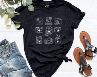 May include: A black short-sleeve t-shirt featuring a white line art graphic of nine different vintage cameras arranged in a grid. The shirt is paired with blue denim jeans and a pair of brown leather thong sandals with black straps.