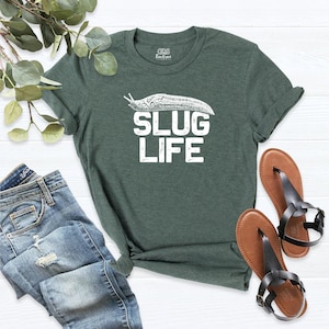 May include: A dark green short-sleeve t-shirt featuring a white graphic of a slug above the text SLUG LIFE in a distressed font. The shirt is displayed alongside blue denim jeans and a pair of brown leather thong sandals.