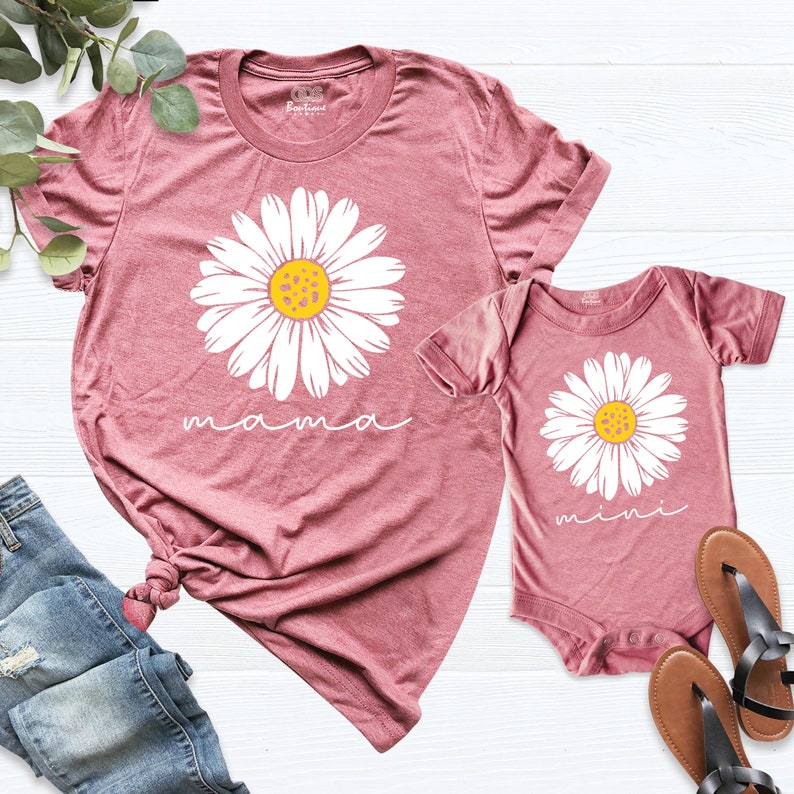 May include: A matching set of two shirts, one for an adult and one for a baby. The adult shirt is a dusty rose color with a white daisy and the word "mama" printed on it. The baby shirt is the same color with a white daisy and the word "mini" printed on it.