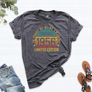 May include: A dark heather gray short-sleeve t-shirt featuring a circular graphic with the text 70 YEARS OF BEING AWESOME, VINTAGE 1956, and LIMITED EDITION in yellow, blue, and orange colors, with a distressed, retro-inspired design style.