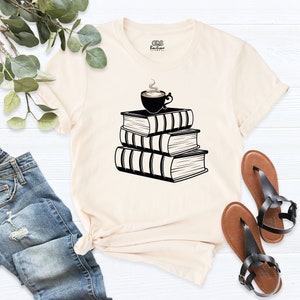 May include: A cream colored t-shirt with a black graphic of a cup of coffee sitting on top of a stack of three books.
