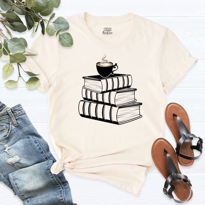May include: A cream colored t-shirt with a black graphic of a cup of coffee sitting on top of a stack of three books.