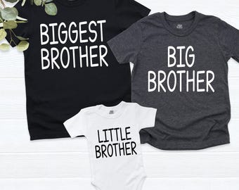 May include: A black t-shirt with white text reading BIGGEST BROTHER, a dark gray t-shirt with white text reading BIG BROTHER, and a white baby bodysuit with black text reading LITTLE BROTHER, all featuring short sleeves and crew necks.