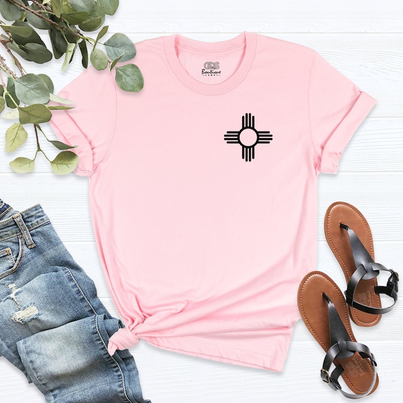 K&ouml;nnte beinhalten: Rosa T-Shirt mit einem schwarzen Zia-Symbol auf der Vorderseite. Das T-Shirt ist aus einem weichen, bequemen Material gefertigt und hat eine lockere Passform.