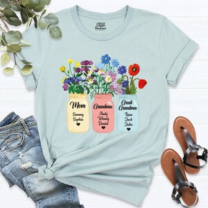 May include: Light blue t-shirt with a floral design featuring three mason jars with the words "Mom", "Grandma", and "Great-Grandma" and personalized names underneath each jar.