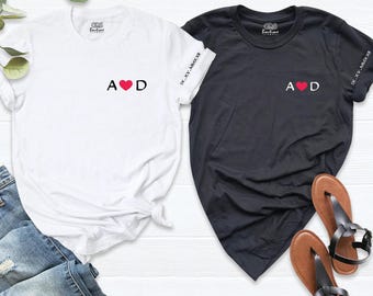May include: A white t-shirt and a black t-shirt, each featuring the letters A and D separated by a red heart on the chest. The left sleeve of each shirt displays the Roman numeral text IX.XV.MMXXII. Both shirts have a round neckline.