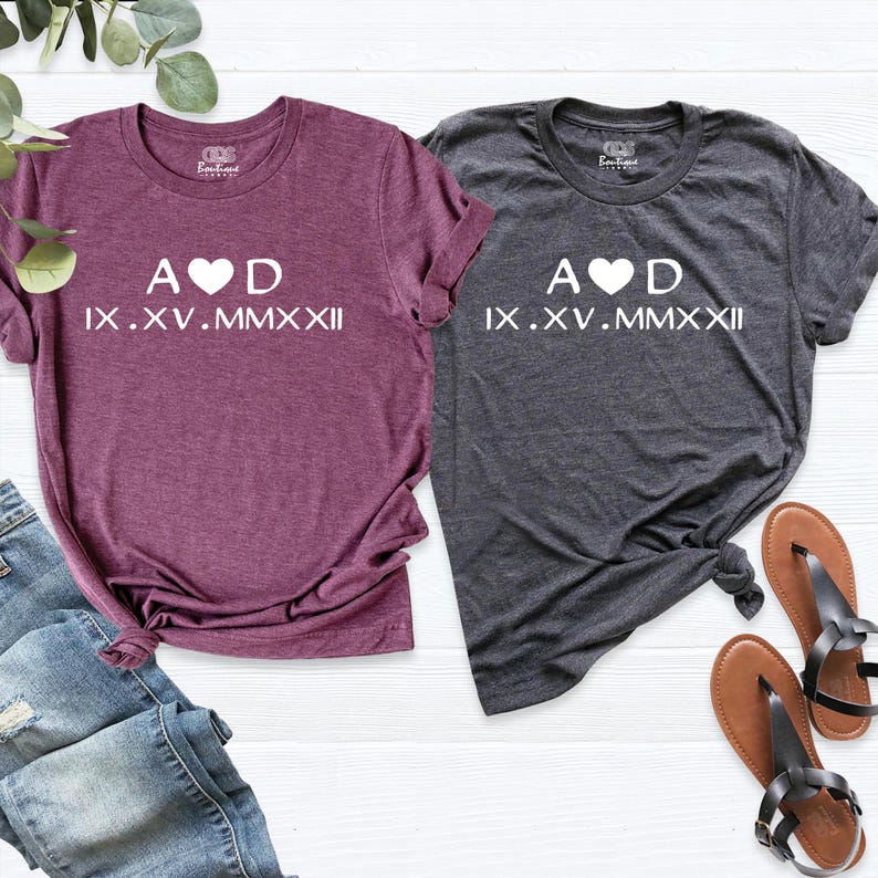 May include: Two t-shirts with a heart and the letters A and D, and the date IX.XV.MMXXII in Roman numerals. The shirts are one maroon and one grey.