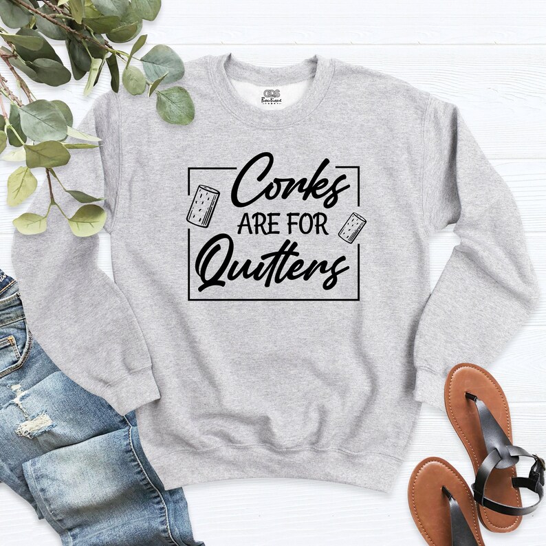 Op de afbeelding: Een grijs sweatshirt met de tekst "Corks are for Quitters" in zwarte letters. Twee kleine kurk afbeeldingen bevinden zich aan beide kanten van de tekst.