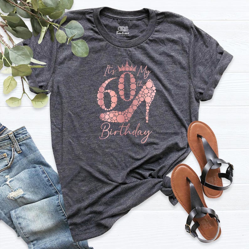 May include: A dark gray t-shirt with a rose gold design that says "It's My 60th Birthday" with a crown and a high heel shoe made of circles.
