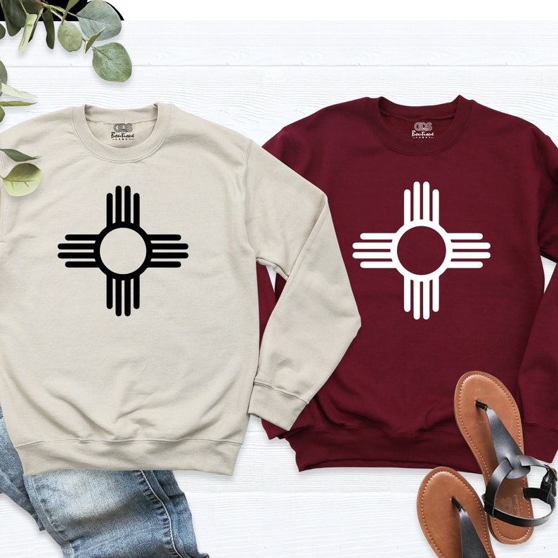 May include: Two long-sleeved sweatshirts with a black and white Zia symbol on a beige and maroon background. The sweatshirts are laid out on a white surface with brown and black sandals.