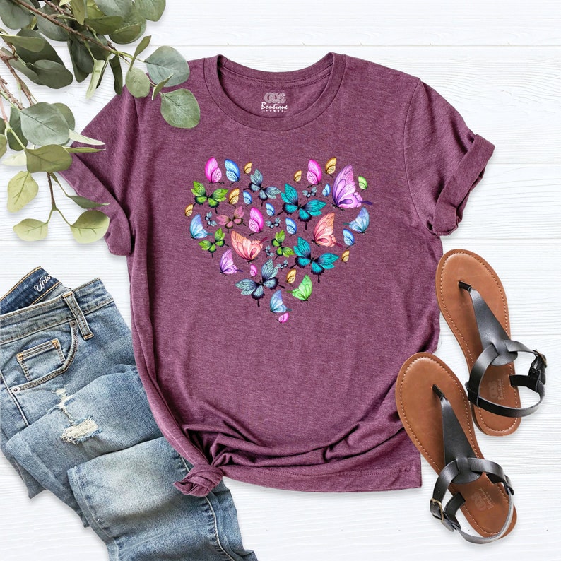 Puede incluir: Una camiseta color granate con un dise&ntilde;o en forma de coraz&oacute;n hecho de mariposas de colores. La camiseta tiene cuello redondo y mangas cortas.