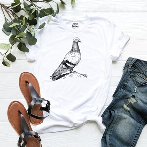 Op de afbeelding: Wit T-shirt met een zwart-witte illustratie van een duif op een tak. Het T-shirt ligt plat op een wit houten oppervlak met een paar bruine sandalen en een blauwe jeans.