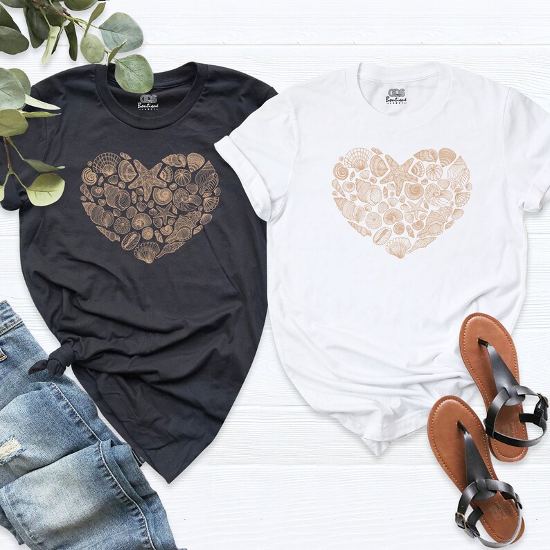 May include: Two t-shirts with a heart-shaped design made of seashells. The t-shirts are black and white. The seashells are a light brown color.