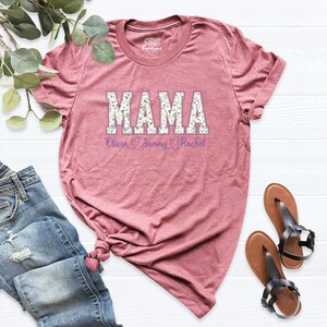 Puede incluir: Una camiseta color malva con un estampado floral de "MAMA" y los nombres Olivia, Sammy y Rachel impresos debajo.