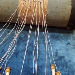 May include: A close-up of a bobbin lace project in progress. White thread is wound around wooden bobbins and strung through a pattern of pins on a pillow. The lace is being created on a wooden pillow with a green fabric cover.