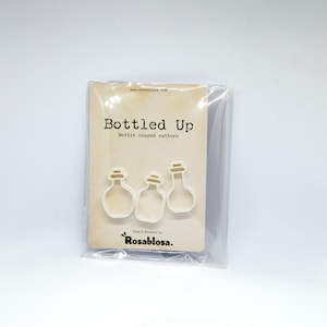 May include: A package of three white plastic bottle-shaped cookie cutters. The package is labeled "Bottled Up" and "Bottle shaped cutters" with the text "Made & designed by Rosablosa."