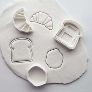 Puede incluir: Cinco cortadores de galletas de plástico blanco sobre una superficie blanca. Los cortadores tienen forma de croissant, rebanada de pan, forma redonda y dos formas diferentes de tostadas.