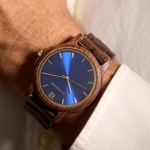 May include: A wooden wristwatch with a blue face and gold-colored hands and accents. The watch has a brown wooden band and is worn on a wrist. The watch face has the text "NEW ZEALAND" and the Roman numeral "IIX".
