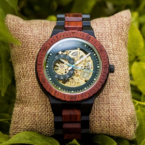May include: A wooden wristwatch with a green face and a red and black wooden band. The watch has a skeleton design, revealing the inner workings of the watch. The watch is on a brown burlap pillow.