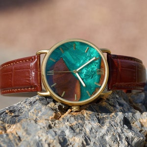 Puede incluir: Un reloj de pulsera dorado con una correa de cuero marr&oacute;n. La esfera del reloj presenta un dise&ntilde;o &uacute;nico con tonos verdes y marrones. Los n&uacute;meros romanos marcan las horas. El reloj se muestra sobre una superficie rocosa texturizada.