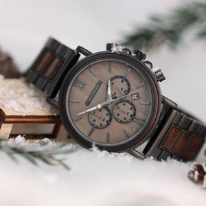 Pu&ograve; includere: Un orologio cronografo in legno marrone con cinturino in metallo nero. Il quadrante dell'orologio ha indici orari in numeri romani e una finestra della data alle 4.