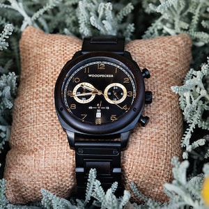 Puede incluir: Un reloj de pulsera de madera negra con una esfera dorada y agujas doradas. El reloj tiene una función de cronógrafo y el nombre de la marca "WOODPECKER" está impreso en la esfera.