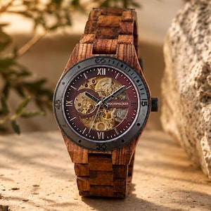 May include: A wooden wristwatch with a brown band and a burgundy face. The watch face has visible gears and Roman numerals. The watch is displayed on a textured stone surface with foliage in the background.