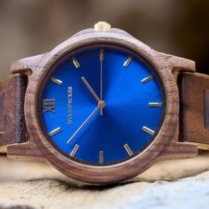 May include: A wooden wristwatch with a blue face and gold-colored hands and markers. The watch has a brown wooden band and a gold-colored crown. The watch face has the word "WOODPICKER" printed on it.