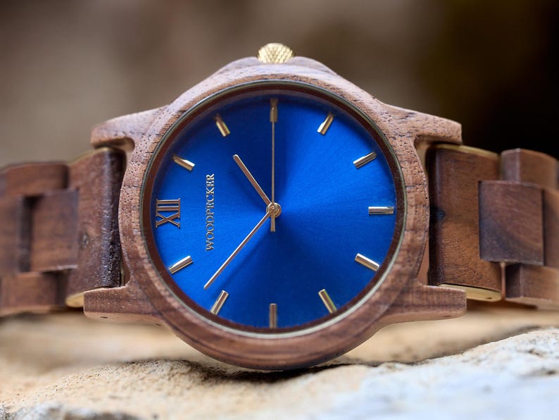 May include: A wooden wristwatch with a blue face and gold-colored hands and markers. The watch has a brown wooden band and a gold-colored crown. The watch face has the word "WOODPICKER" printed on it.