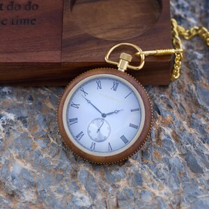 May include: A gold pocket watch with Roman numeral markings on a white face. The watch has a brown wooden case and is attached to a gold chain.