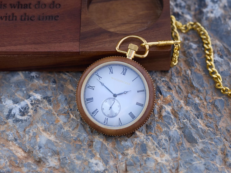 May include: A gold pocket watch with Roman numeral markings on a white face. The watch has a brown wooden case and is attached to a gold chain.