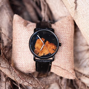 Puede incluir: Un reloj de pulsera negro con una correa de cuero y una esfera de madera &uacute;nica. El reloj se muestra sobre un coj&iacute;n beige, con un fondo natural de ramas de &aacute;rboles y tela.