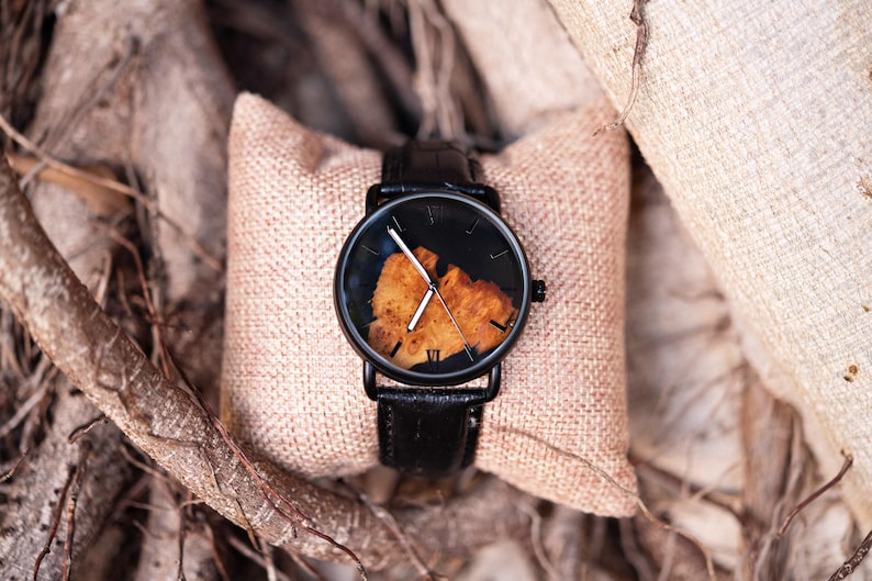 Puede incluir: Un reloj de pulsera negro con una correa de cuero y una esfera de madera &uacute;nica. El reloj se muestra sobre un coj&iacute;n beige, con un fondo natural de ramas de &aacute;rboles y tela.