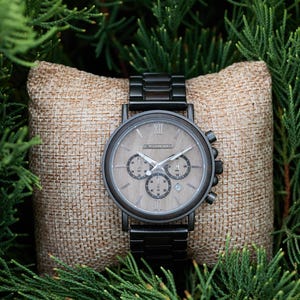 Pu&ograve; includere: Un orologio da polso cronografo nero con quadrante in legno e cinturino in metallo nero. L'orologio &egrave; su un cuscino di juta marrone.