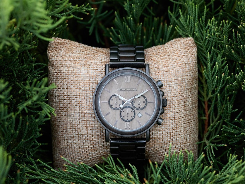 Pu&ograve; includere: Un orologio da polso cronografo nero con quadrante in legno e cinturino in metallo nero. L'orologio &egrave; su un cuscino di juta marrone.