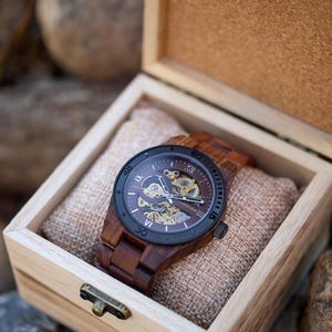 Puede incluir: Un reloj de madera con una esfera negra y n&uacute;meros romanos. El reloj est&aacute; en una caja de madera con un forro de tela marr&oacute;n.