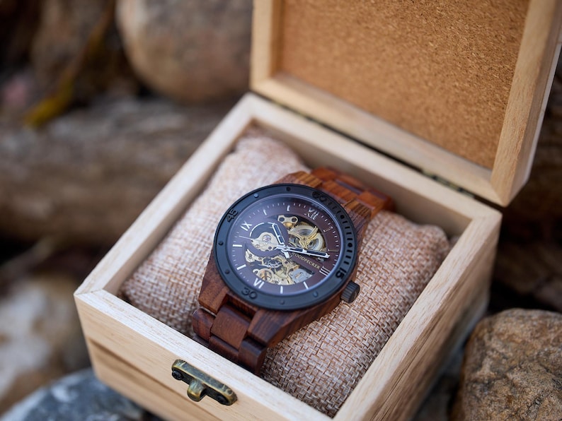 Puede incluir: Un reloj de madera con una esfera negra y n&uacute;meros romanos. El reloj est&aacute; en una caja de madera con un forro de tela marr&oacute;n.