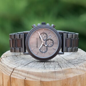 Puede incluir: Un reloj de pulsera cron&oacute;grafo de madera marr&oacute;n con una banda de metal negro. La esfera del reloj tiene marcadores de hora en n&uacute;meros romanos y una ventana de fecha.