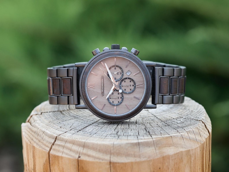 Puede incluir: Un reloj de pulsera cron&oacute;grafo de madera marr&oacute;n con una banda de metal negro. La esfera del reloj tiene marcadores de hora en n&uacute;meros romanos y una ventana de fecha.